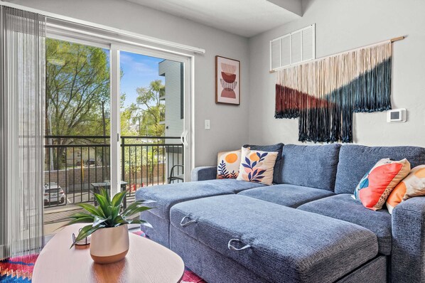 A modern living room with a grey sofa, a small table with a plant, and a view of the outside through a sliding glass door. - Downtown SLC 2BR Delta & Conference (Salt Lake City)