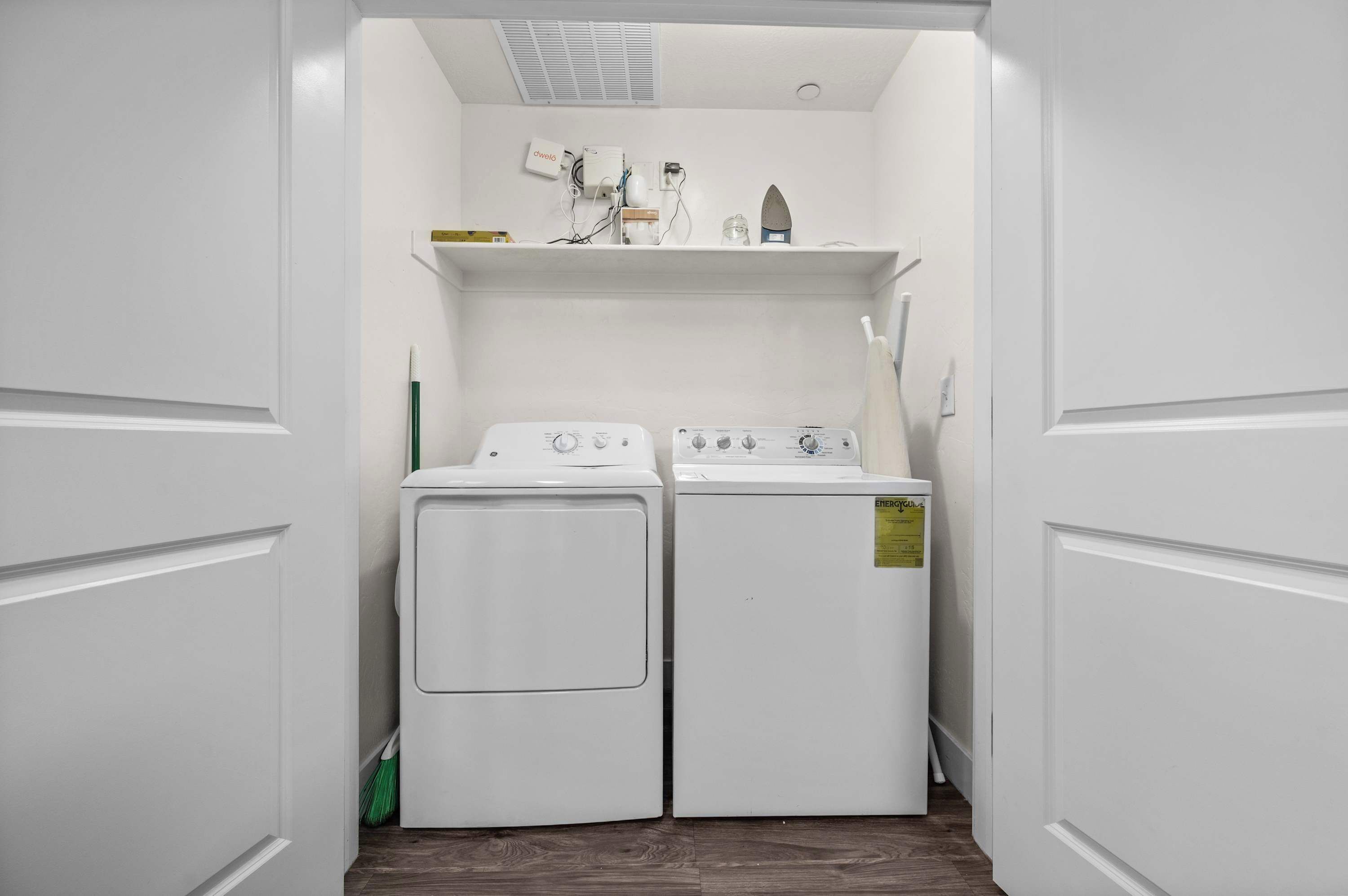 A laundry room with two white washing machines and a dryer, a shelf with various items, and a vent on the ceiling.