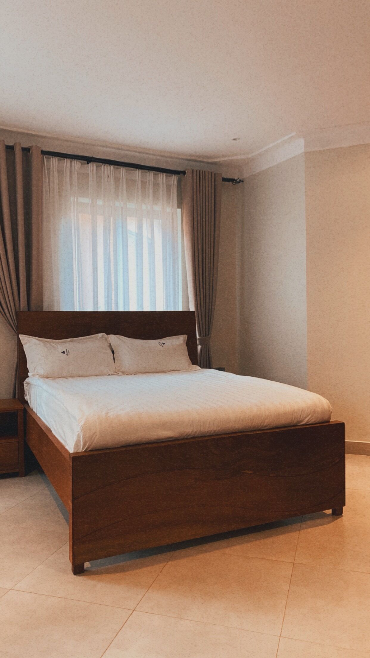 Une chambre à coucher avec un lit en bois, une table de chevet, une fenêtre avec des rideaux et un mur uni.