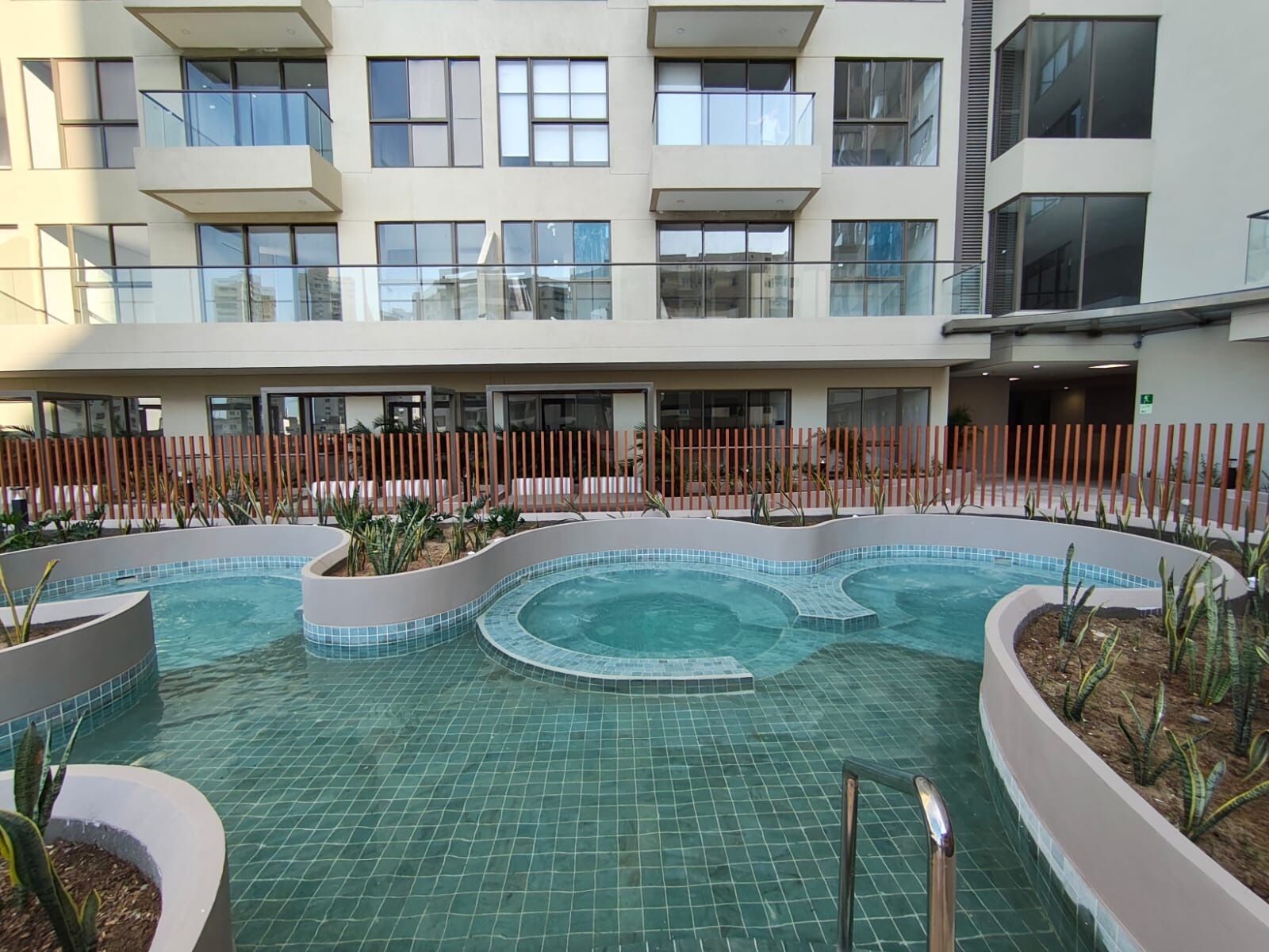 Un edificio moderno con piscina, varios balcones y un área cercada con plantas.