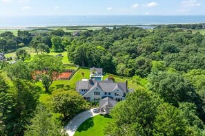 A large house surrounded by dense greenery, a curved driveway, and a view of the ocean.