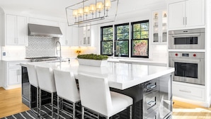 A modern kitchen with white cabinetry, a central island with chairs, a built-in oven, and a large window with a view of trees. - Luxurious expansive villa in prestigious Watch Hill set on 5 acres (Westerly)