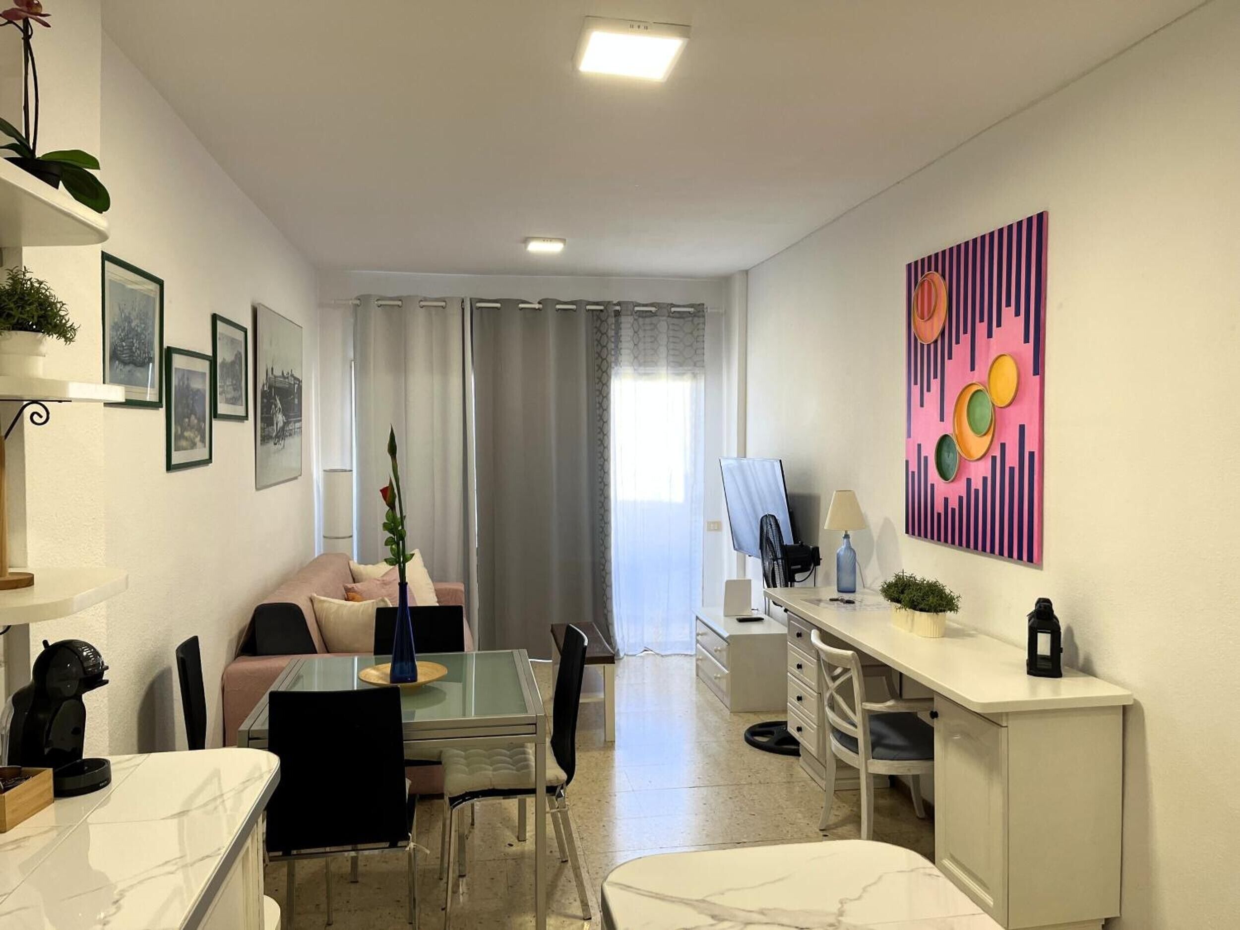 A modern living room with a glass dining table, a sofa, a desk with a computer, and a large abstract painting on the wall.