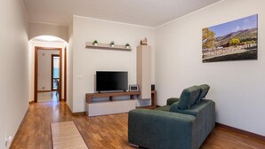 Un salón con televisor de pantalla plana, un sofá, piso de madera, un cuadro en la pared y un pasillo que da a otra habitación.