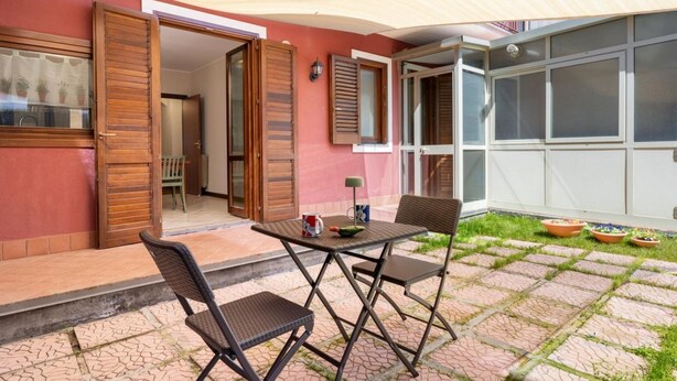 Un patio al aire libre con mesa y sillas, un área cubierta y vista del interior a través de puertas abiertas.