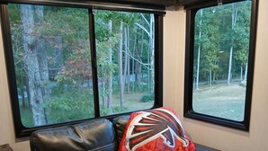 Ein schwarzes Ledersofa mit einem rot-schwarzen Kissen in einem Zimmer mit zwei Fenstern mit Blick auf ein bewaldetes Gebiet.