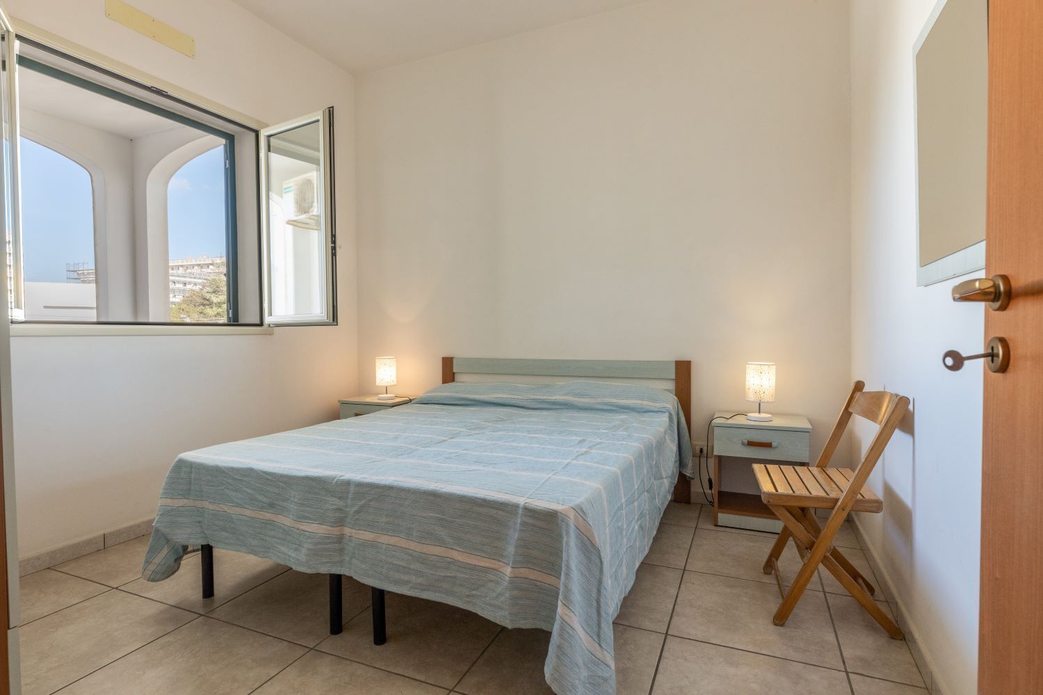 Un dormitorio con cama, mesitas de noche, una silla y una ventana.