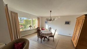 A spacious room with a dining table, chairs, a chandelier, a radiator, and a large window with curtains. - Apartment Hoeri with Private Garden, Terrace, and Wi-Fi (Uhldingen-Mühlhofen)