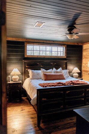 Ein Schlafzimmer mit einem Holz-Kopfteil, einem großen Fenster, zwei Nachttischen mit Lampen sowie einem Bett mit Kissen und Decke.