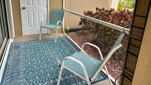 Een overdekte buitenruimte met een deur, twee stoelen, een kamerkleed met patroon en uitzicht op een tuin met planten en een hek.