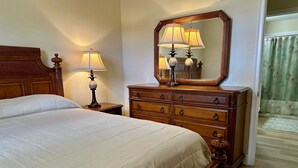 Een net opgemaakt bed met een houten hoofdbord, een houten dressoir met lades, een grote spiegel en een bedlampje.