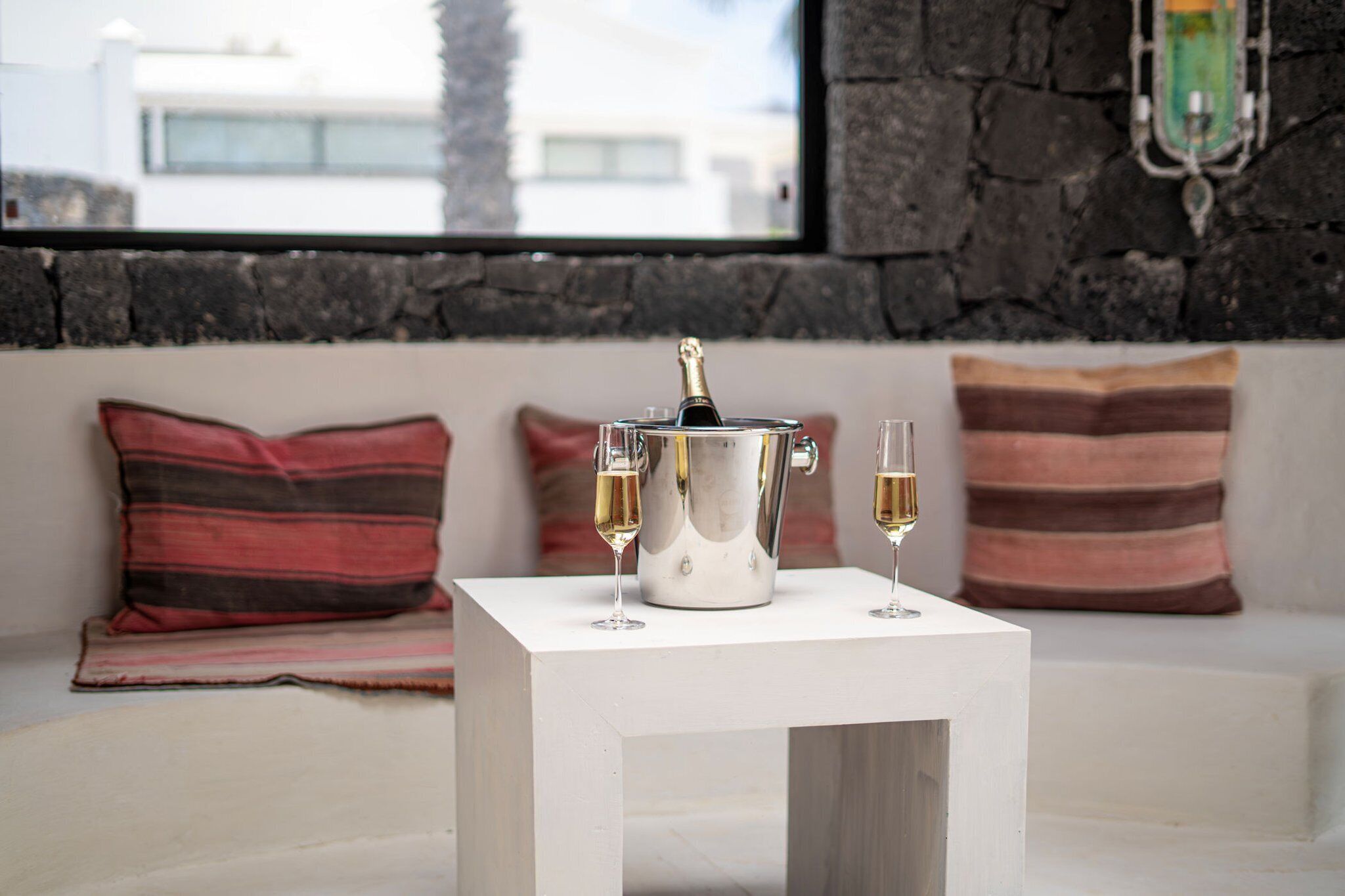A white table with a champagne bucket and two glasses of champagne on a white sofa with striped pillows.