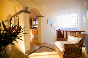 Ein Wohnzimmer mit einem Korbstuhl, einer Küche im Hintergrund und Blick durch ein Fenster.