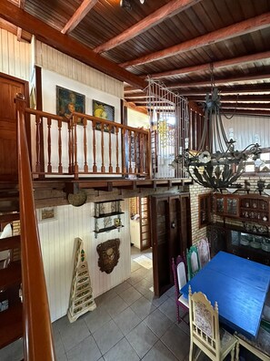 A spacious room with a high ceiling, wooden beams, a staircase, a chandelier, and a dining area with a blue table.