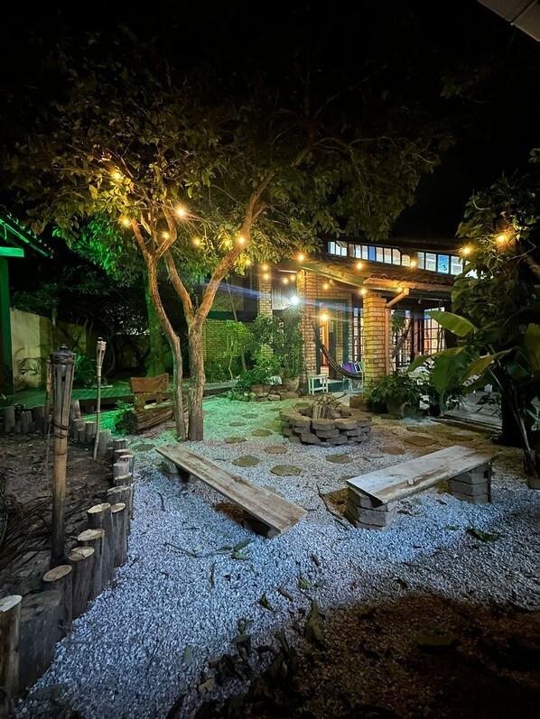 A nighttime outdoor area with a brick building, string lights, wooden benches, and a gravel path.
