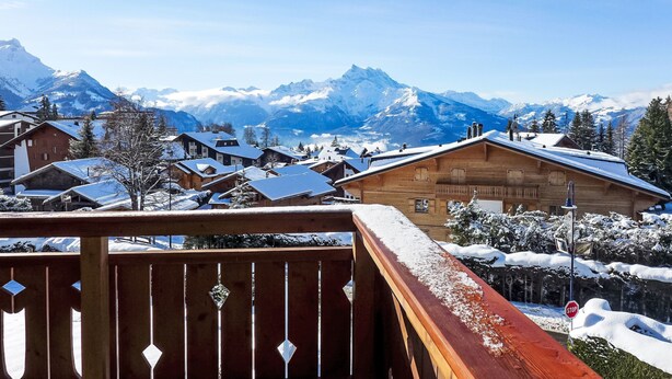 Een houten chalet met een balkon met uitzicht op een besneeuwde bergdorp en een helderblauwe lucht.