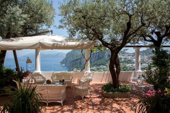 Eine Terrasse mit Korbmöbeln, einem Sonnensegel und Blick auf Meer und Berge.