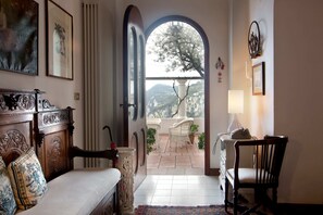 Ein Zimmer mit Bett, Sessel, Schreibtisch und Blick durch eine Bogenöffnung auf einen Balkon mit Pflanzen und einem Bergpanorama.