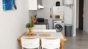 Una cuina compacta amb microones, bullidor i rentadora, taula de menjador amb caires i un quadre abstracte blau a la paret.