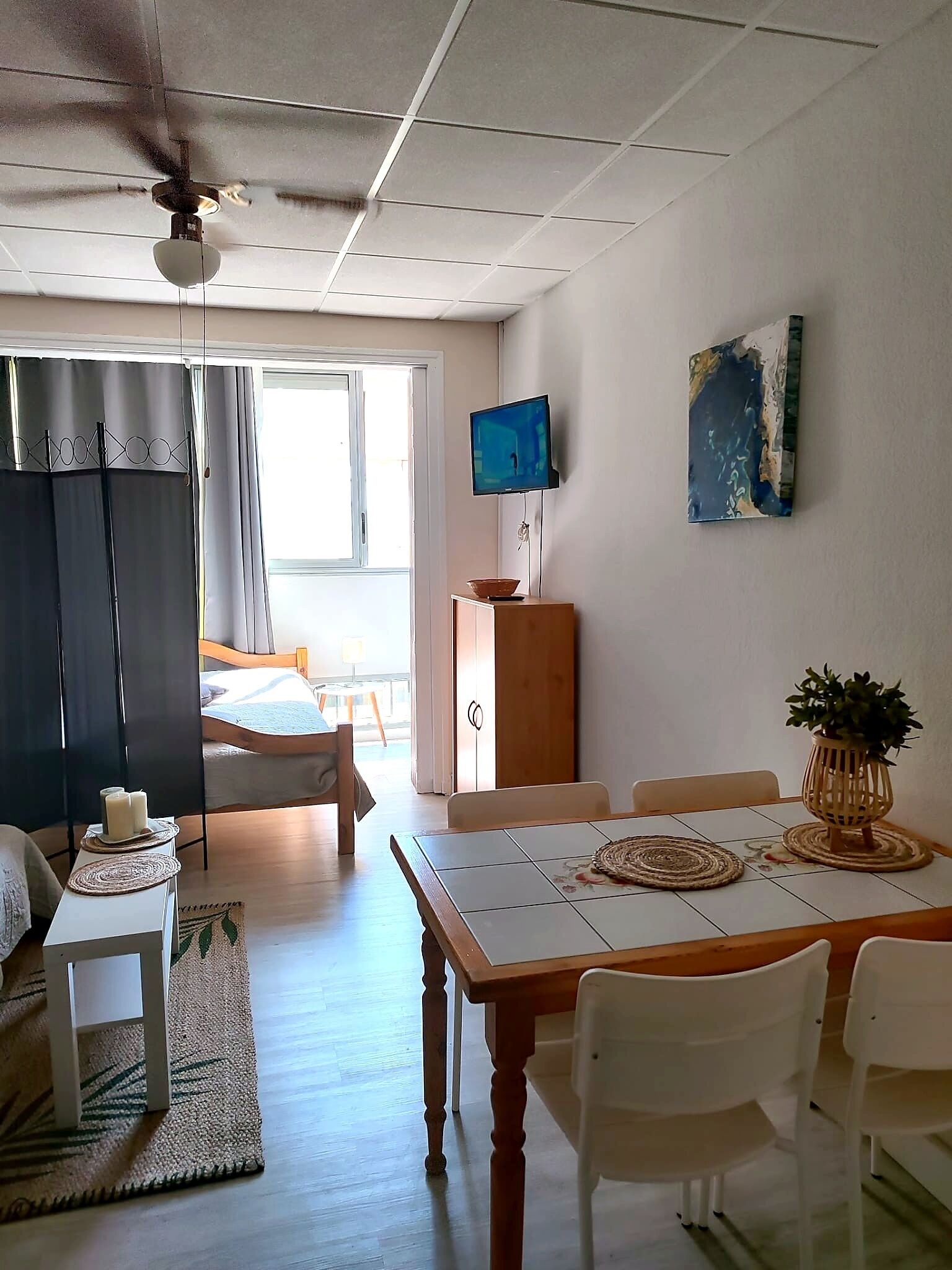 Une pièce comprenant un lit, un téléviseur, une table à manger avec des chaises, un ventilateur de plafond et une fenêtre.