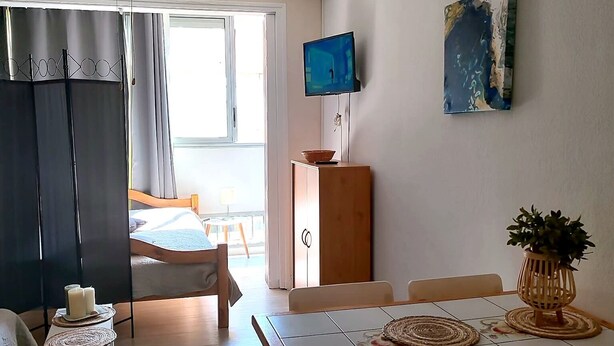 Una habitació amb un llit, una televisió, una taula de dinar amb cadires, un ventilador de sostre i una finestra.