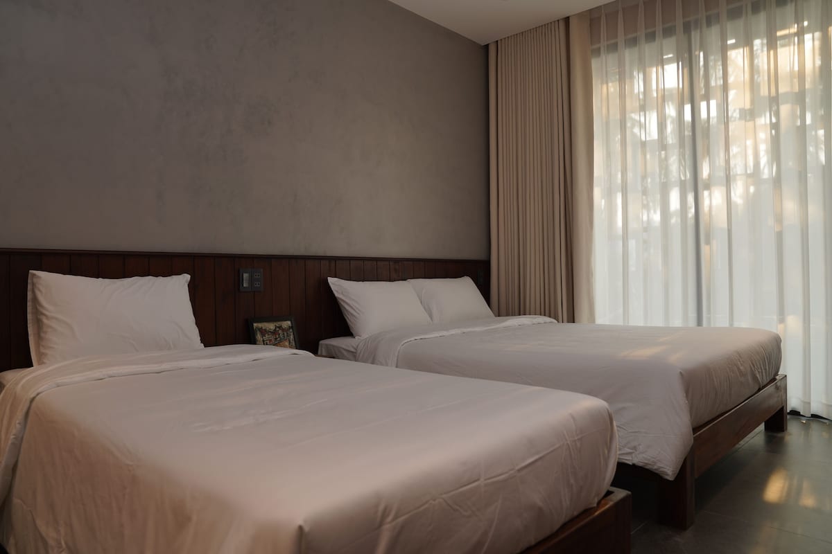 Camera d'albergo con due letti, una testiera in legno, un'ampia finestra con tende leggere e una parete spoglia.