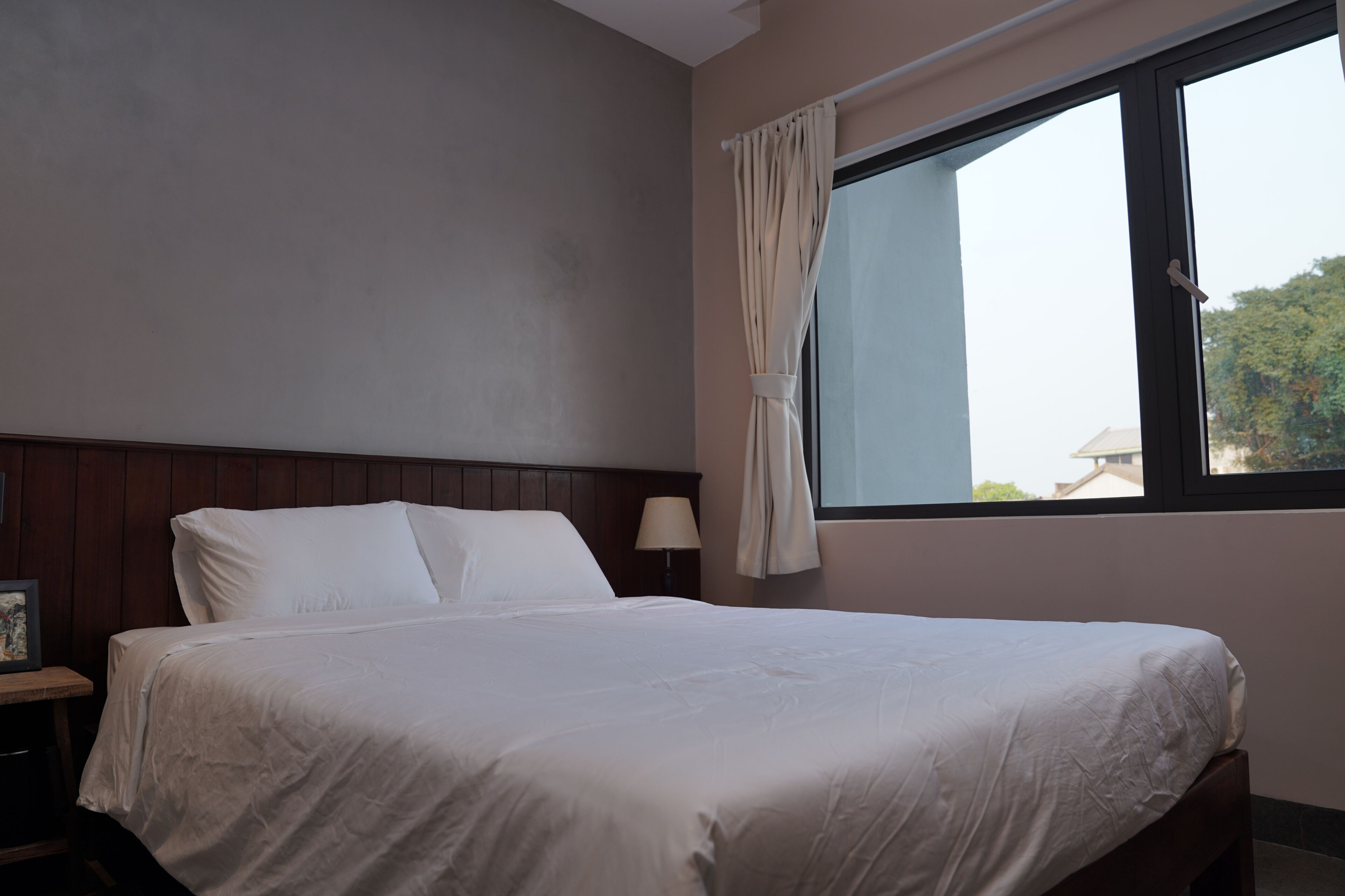 A neatly made bed in a room with a window, a nightstand with a lamp, and a wooden headboard.