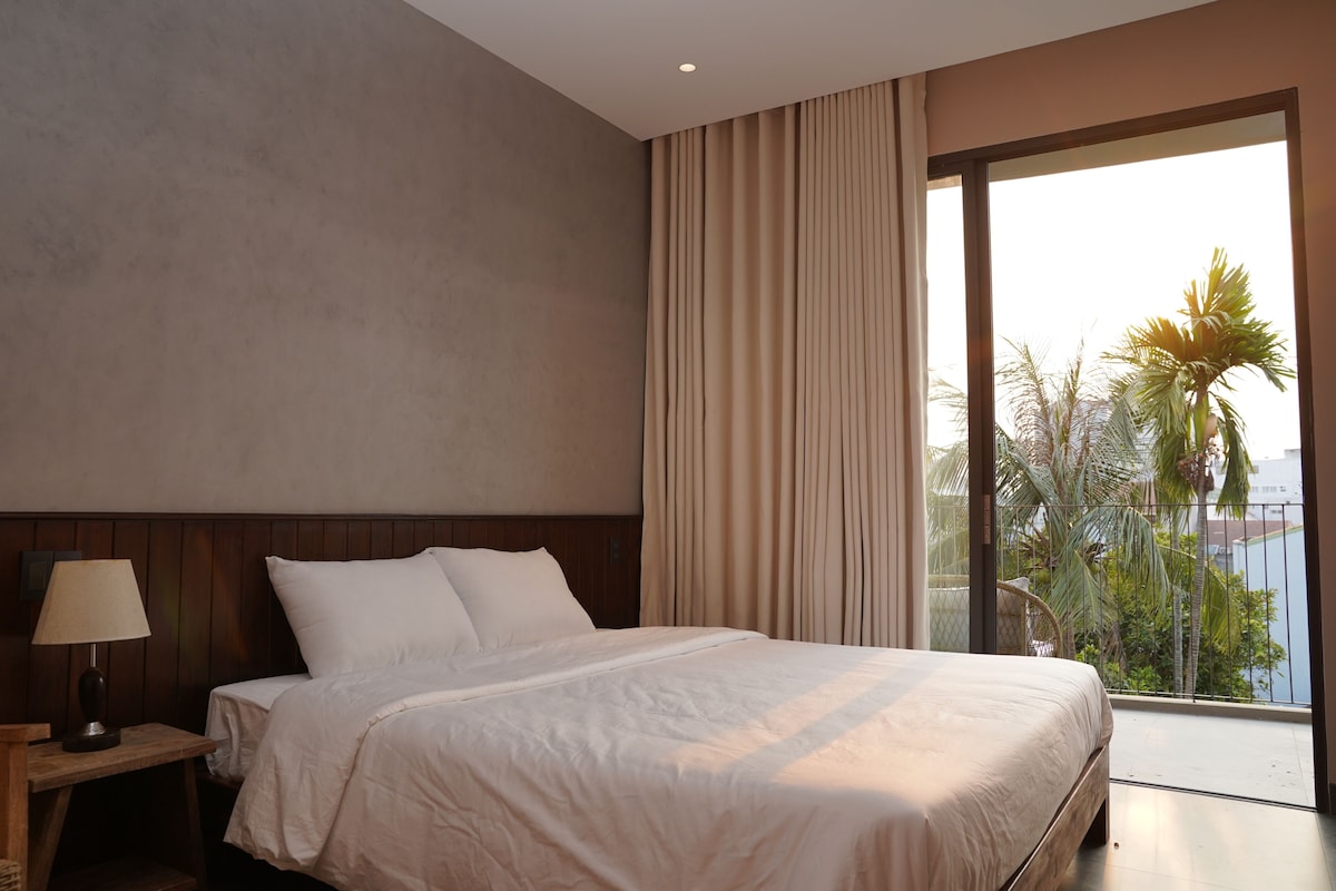 Camera d'albergo con un letto grande, un comodino con una lampada e vista su palme attraverso una finestra.
