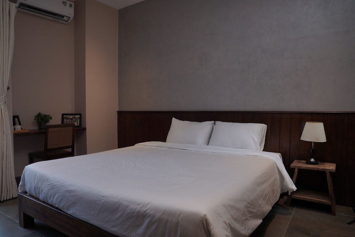 Un letto rifatto con cura in una camera d'albergo, con una testiera in legno, un comodino con una lampada e una piccola pianta.
