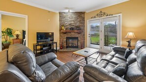A living room with a stone fireplace, leather sofas, a television, and a balcony with a view. - Baskins Creek 212 🐻 Sleeps 6 | Pool Views, Hot Tub & Stone Fireplace 🔥 (Gatlinburg)