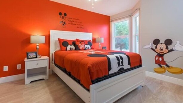 Une chambre avec un lit blanc, une literie rouge, une fresque murale représentant Mickey Mouse, une table de chevet et une fenêtre.