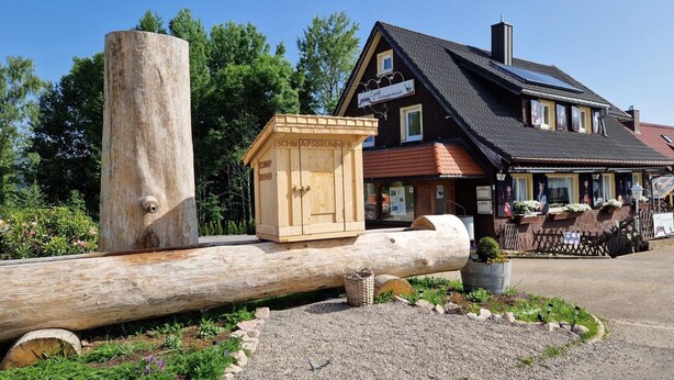 A rustic wooden structure with a sign, a large log, and a building with a sloped roof and solar panels. - Apartment 'Fewo Am Adlerweiher' with Shared Terrace, Shared Garden and Wi-Fi (Feldberg (Schwarzwald))