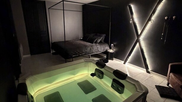 A modern bedroom with a large bathtub, a four-poster bed, and a wall-mounted X-shaped light fixture. - The secret lodge (Dolus-le-Sec)