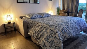 A modern bedroom with a large bed, a bedside table with a lamp, a wall-mounted mirror, and a view of the outdoors through a window with curtains. - L'Eden in Downtown Saint-Tropez with a Pool (Saint-Tropez)