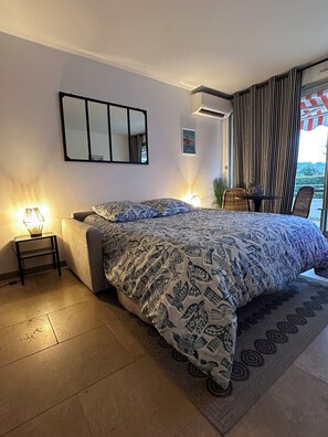 A modern bedroom with a large bed, a bedside table with a lamp, a wall-mounted mirror, and a view of the outdoors through a window with curtains. - L'Eden in Downtown Saint-Tropez with a Pool (Saint-Tropez)