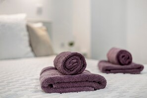 A neatly folded purple towel on a white textured bedspread in a room with a white wall and a white pillow in the background. - Apartment 'Casa Pereira 6' with Wi-Fi (Ponta do Pargo)