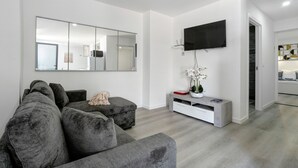Ein modernes Wohnzimmer mit einem grauen Ecksofa, einem flachbildfernseher an der Wand, einem weißen Konsolentisch mit einer Blumenvase und einem Flur, der zu einem anderen Raum führt.