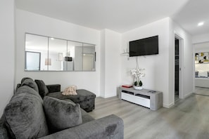 Ein modernes Wohnzimmer mit einem grauen Ecksofa, einem flachbildfernseher an der Wand, einem weißen Konsolentisch mit einer Blumenvase und einem Flur, der zu einem anderen Raum führt.