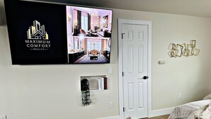 A modern living room with a flat-screen TV displaying a real estate video, a white door, and a wall-mounted decorative piece. - Designer Luxe Studio | King Suite + Work Space (Sacramento)