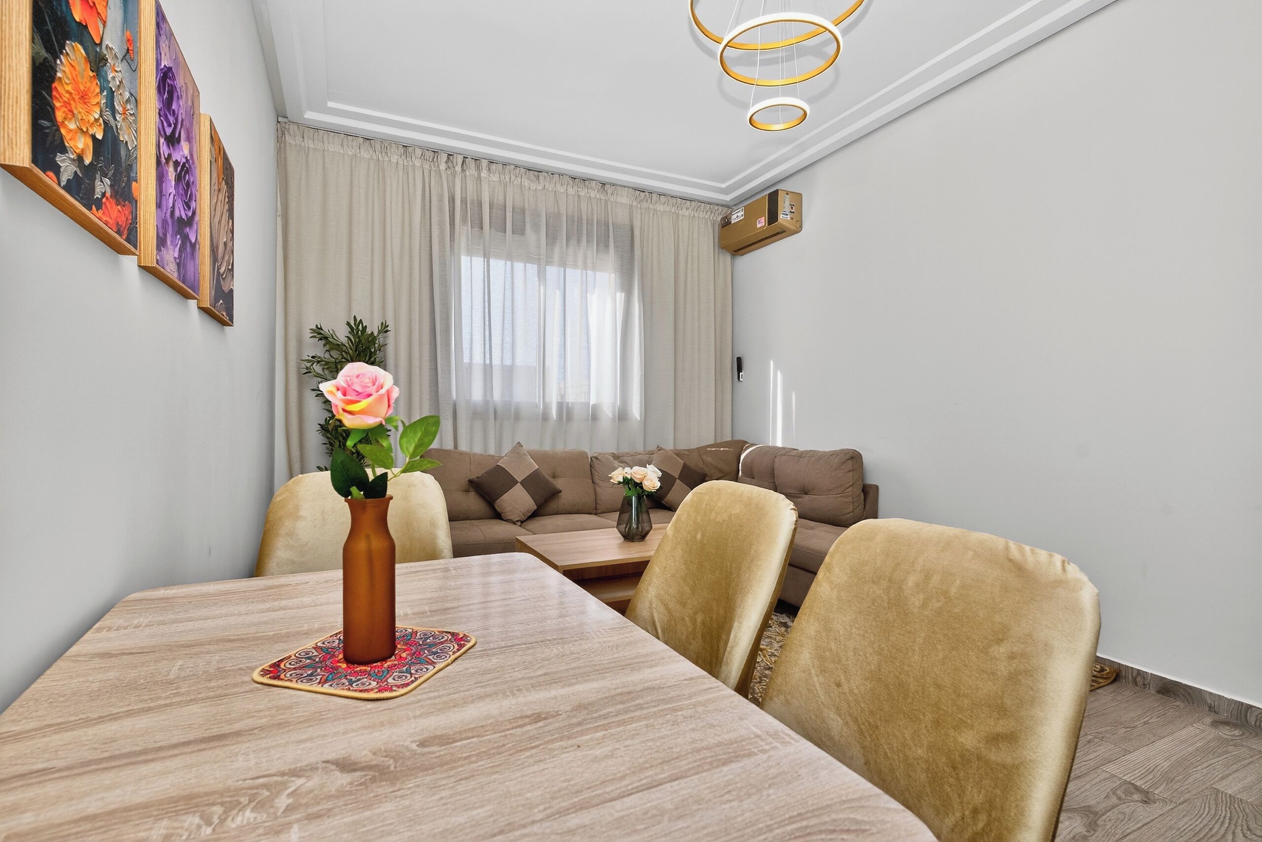 A dining area with a wooden table, chairs, a vase with flowers, a sofa, and a window with curtains.
