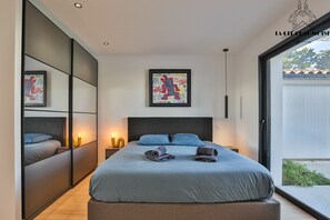 A modern bedroom with a large bed, a side table with a lamp, a framed artwork above the bed, and a sliding door leading to a balcony. - La Villa Pop I Talmont I 12 pers I Piscine & Pétanque (Talmont-Saint-Hilaire)