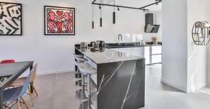 Moderni keittiö, jossa on keskeinen saareke, baarituolit, ruokapöytä tuoleineen sekä taideteoksia seinillä.