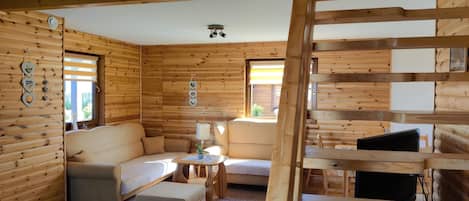 Interior de una cabaña de troncos con sofá, sillón, reposapiés y televisor sobre un soporte de madera.