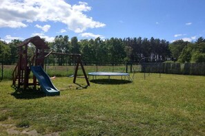 Un'area giochi con altalene, scivolo e trampolino, immersa nel verde con alberi sullo sfondo.