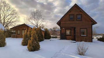 Una cabaña de madera con techo a dos aguas, rodeada de terreno cubierto de nieve y árboles escasos.