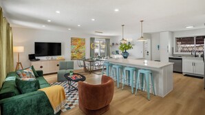 A modern living room with a sofa, a coffee table, and a TV. There is a kitchen area with a dining table and bar stools. - Renovated retreat in NE Edmonton with a sauna, Pac-Man & Street Fighter arcade, home theater with projector & popcorn machine. 16 min to downtown, 8 min to Rundle Park golf. Built for families, friend groups & celebrations up to 13 guests. (Edmonton)