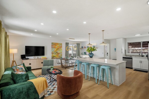 A modern living room with a sofa, a coffee table, and a TV. There is a kitchen area with a dining table and bar stools.