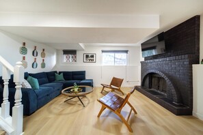 A living room with a dark brick fireplace, a blue sofa, a wooden coffee table, and a wooden chair. - Older home in NE Edmonton with a renovated retro interior: hot tub, sauna, Pac-Man & Street Fighter arcade, home theater with projector & popcorn machine. 16 min to downtown, 8 min to Rundle Park golf. Pool closed for the season. Up to 12 guests. (Edmonton)