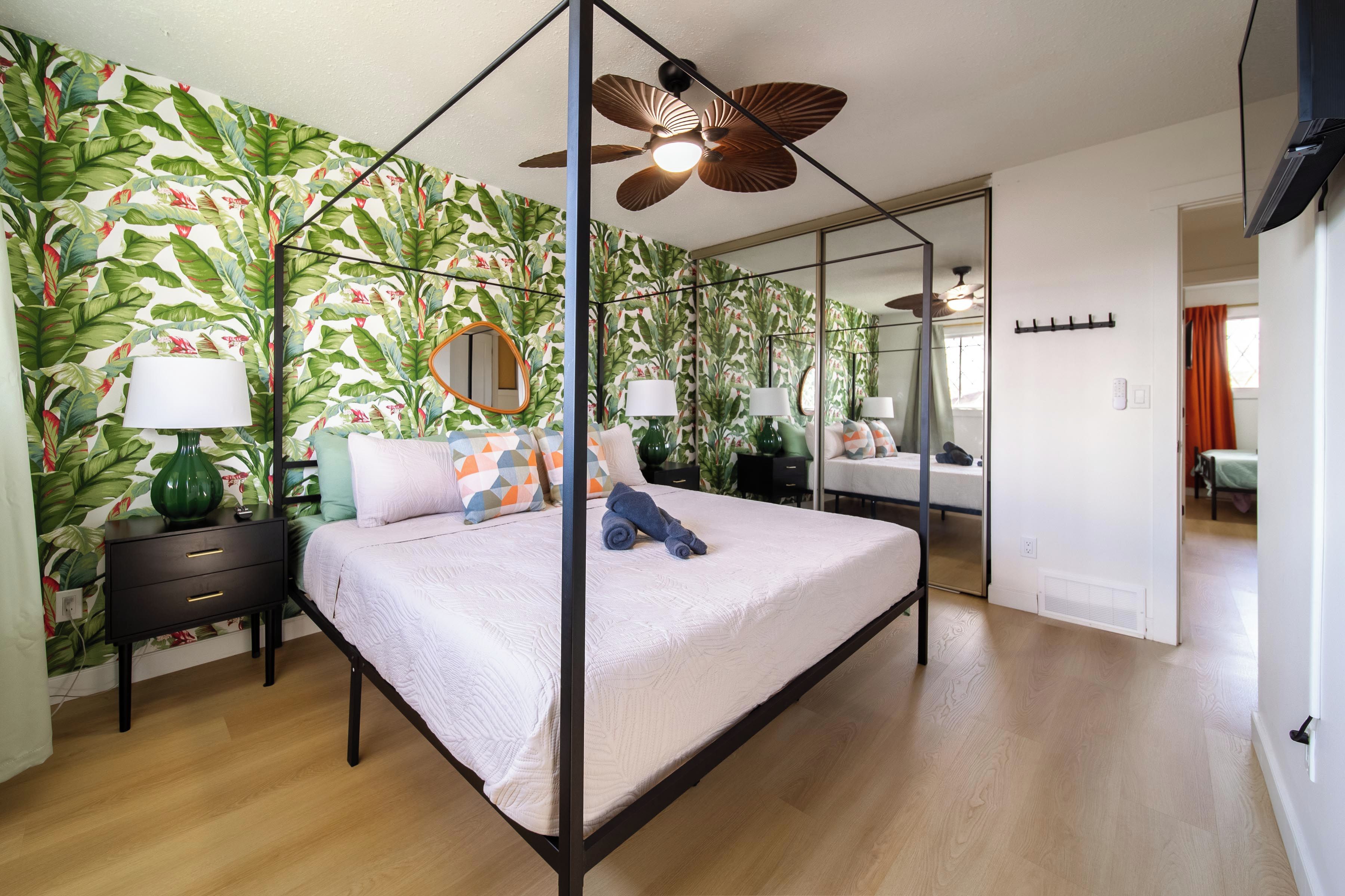 A bedroom with a canopy bed, a ceiling fan, a mirror, and a tropical wallpaper.