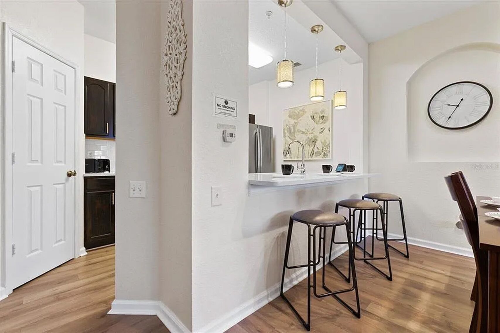 A kitchen with a white countertop, a sink, and three bar stools. There is a clock on the wall and a 'No Smoking' sign.